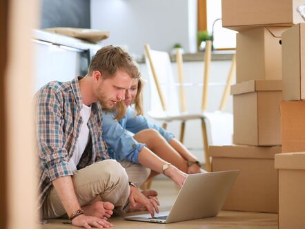 A couple sitting in front of a laptop surrounded by cardboard boxes